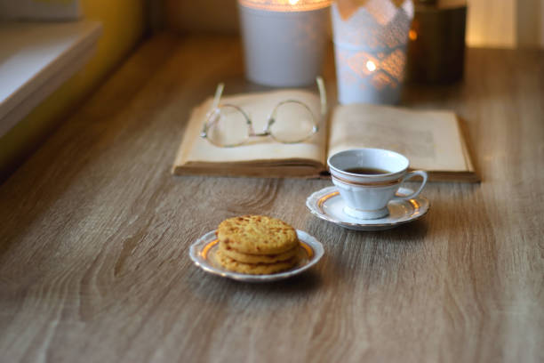 Plate with peanut butter cookies, cup of tea or coffee, open book, reading glasses and lit candles on the table. Selective focus.