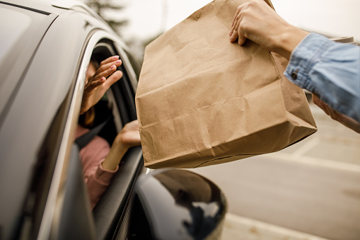 Close up shot of unrecognizable customer sitting in her car, in driver's seat, receiving a coffee and takeaway food order from a service person at the drive through.