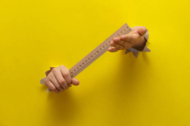 istock Two children's hands stretched through a torn sheet of yellow paper hold the student's ruler 1444436457