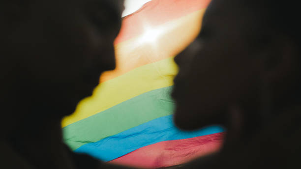 LGBT pride celebration. Queer people of color standing in a romantic embrace in front of a rainbow flag Rainbow flag floating on wind behind two people of african ethnicity standing closely in romantic embrace. LGBT equality pride parade bi black stock pictures, royalty-free photos & images