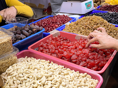 Stock photo showing close-up, elevated view of market stall display of piles of dried fruits and nuts in plastic containers, including dried dates, candied cherries and cashew nuts. Raw cashews are actually unsafe to eat due to containing urushiol which is toxic and can cause skin irritation. Therefore, 'raw' cashews sold in stores have actually been cooked to remove the urushiol and once this is done cashews are considered to be a very healthy snack food being low in sugar but high in fibre as well as a good source of copper, magnesium, and manganese.
