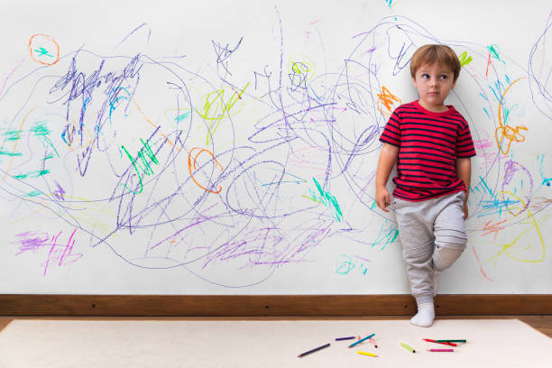 Child mischief. Boy with a distracted face because he drew the entire wall. Child mischief. Boy with a distracted face because he drew the entire wall. Little boy leaning against the white wall where he made many drawings with colored pencils. Kid indoors, at home. child behaving badly stock pictures, royalty-free photos & images