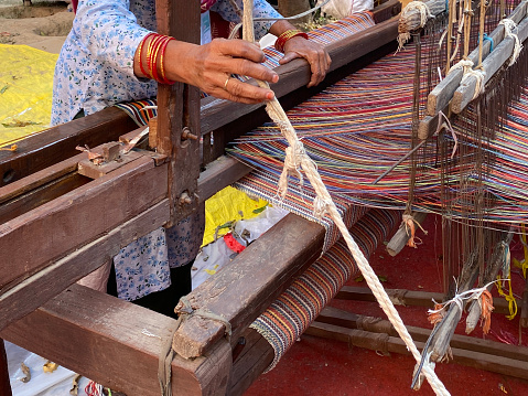 Stock photo showing close-up view of a wooden loom being used by an unrecognisable Indian woman to weave a striped rug.