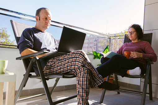 young man working with his laptop sitting on the terrace of his house accompanied by a young woman reading a book and having a coffee or tea on a sunny day.