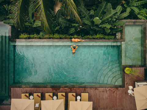 Aerial shot of woman relaxing at the poolside with man in swimming pool. Couple on holiday at luxury resort.