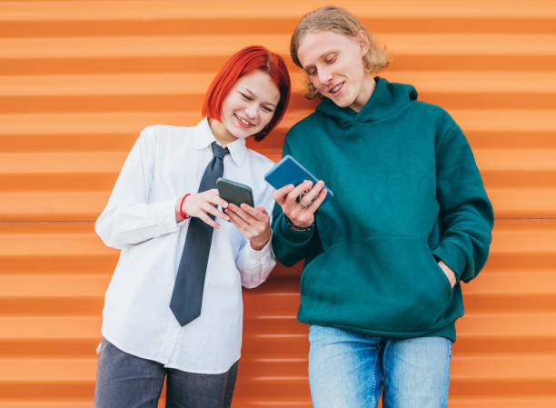 Two caucasian teen friends boy and a girl browsing their smartphone devices while they lean on the orange wall background. Careless young teenhood time and modern technology concept image. dos amigos adolescentes caucásicos niño y una niña navegando por sus dispositivos de teléfono inteligente mientras se apoyan en el fondo naranja de la pared. tiempo de adolescencia joven descuidado e imagen de concepto de tecnología moderna.