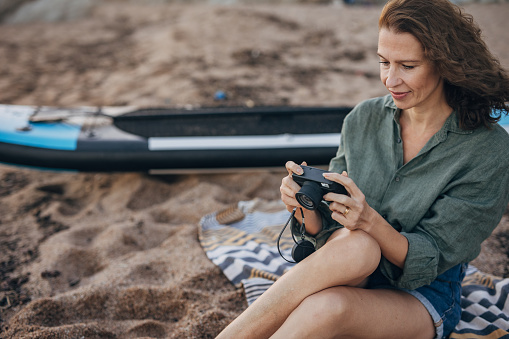 One woman, mature female sitting on the beach in sunset, she is taking pictures with camera.