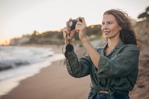 One woman, mature female on the beach in sunset, she is taking pictures with camera.