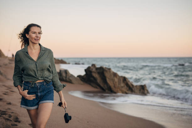 One woman, mature female walking on the beach in sunset, she is holding a camera.