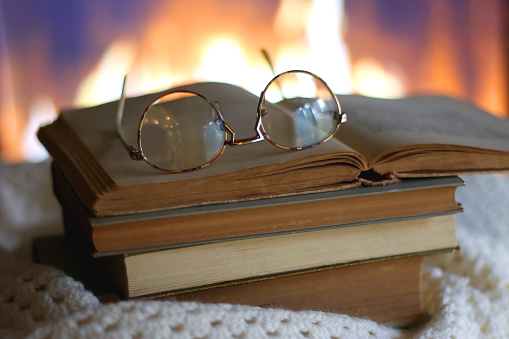 Stack of books, soft blanket, books and reading glasses on the table in front of a fireplace. Selective focus.