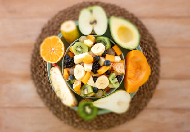 Above view of a fresh inviting fruit salad in a green bowl. Half-cutted fruit around. Healthy eating and wellness concept. Wooden background