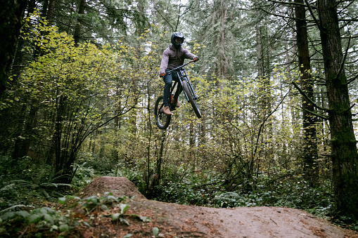 An African American mountain bike rider enjoys an overcast ride through forest trails in Washington state, USA.  He soars through the air on his bike off of a dirt jump.