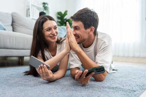Caucasian attractive couple using smartphone play game in living room. Attractive male and female feeling happy and relax, spend leisure time lying down on floor play mobile gaming together in house. stock photo