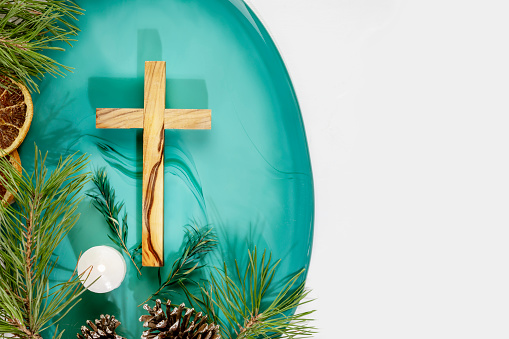 Wood cross laying on a green marble podium with fir branches, cones, candle and dry oranges on a white background with copy space. Christmas holidays. Christian religion card. Flat lay