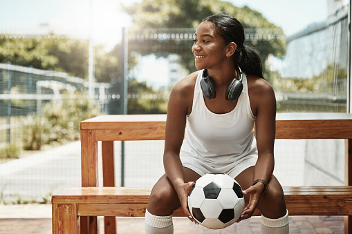 Sports, soccer and a woman on bench with ball in city park in Sao Paulo. Fitness, fun and a happy black woman in Brazil sitting, holding soccer ball and watching a game in summer sun with headphones