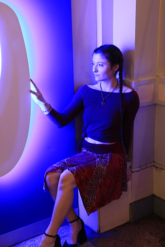 A Caucasian woman sitting next to a multicolored light of blues and greens. She is wearing a necklace, bracelet, blue sweater. patterned skirt and high heels.