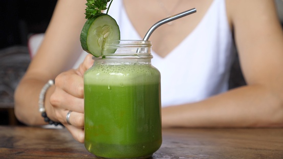 A graceful female hand takes a stylish jar by the handle, scrolling it around the axis with cold-pressed organic green juice with a stable metal drinking straw.