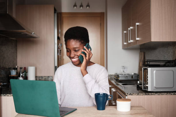 mulher negra sorridente falando no smartphone enquanto usa o laptop - working at home domestic kitchen laptop computer - fotografias e filmes do acervo