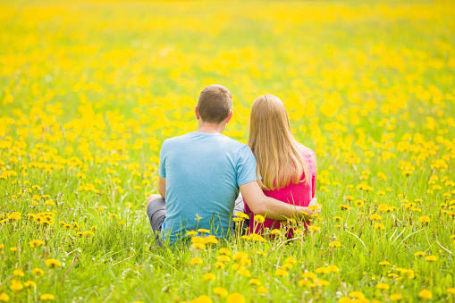 Young adult couple hugging each other and sitting at meadow of fresh yellow blooming dandelions in sunny spring day. Looking far away. Peaceful atmosphere in nature. Back view. Romantic lovely moment.