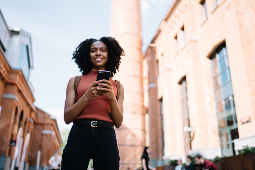 View from below of positive smiling black curly haired woman in casual wear using phone chatting with friend strolling in street looking at camera