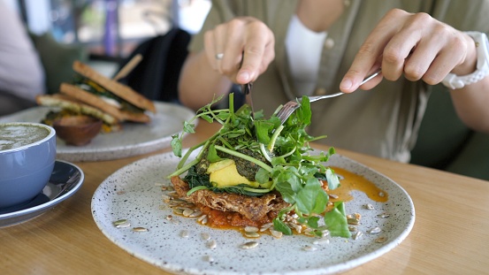 Close-up of a woman cutting a vegan sandwich with cutlery with lots of vegetables and herbs. Then she skewers a slice of the vegetable sandwich on her fork. Plant-based lunch options.