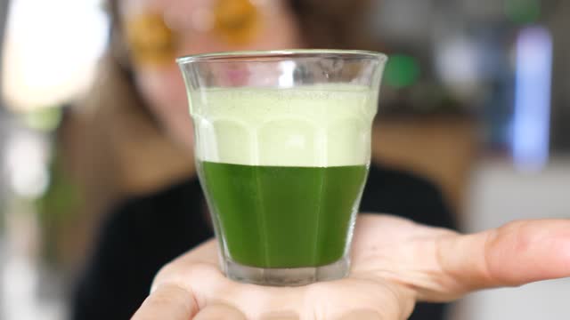 A woman in glasses on a blurred background points to the camera on her palm, a small glass filled with a drink with organic green spirulina. The drink is a storehouse of vitamins, minerals, nutrients.