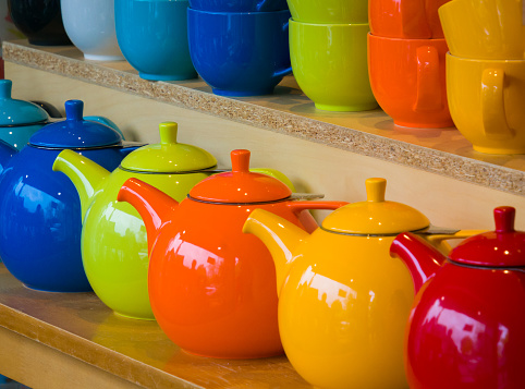 A row of colorful ceramic teapots  rest on a shelf below a row of stacked cups in a shop window.