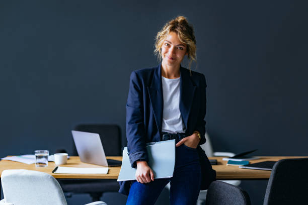 Portrait Of Happy Business Woman Standing In The Office Smiling businesswoman holding files while leaning on a desk and looking at camera. chairperson stock pictures, royalty-free photos & images