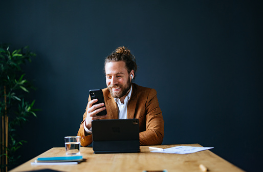 Smiling business man with wireless earphones listening to music on his smartphone while sitting at office desk with a digital tablet, documents and glass of water.