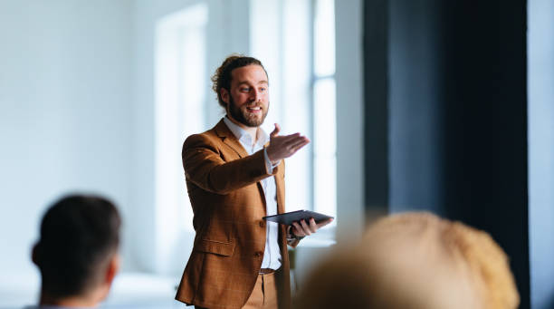 Smiling Business Man Talking And Pointing To Audience During A Q And A Session Happy presenter holding a digital tablet and speaking to unrecognizable multi-ethnic businesspeople at their company. chairperson stock pictures, royalty-free photos & images