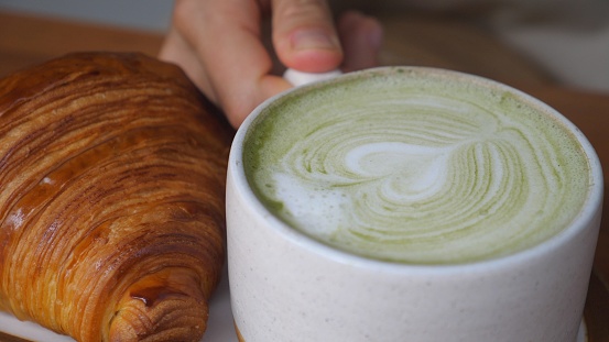 Matcha latte mug decorated with palm leaf shaped milk foam, served with fresh croissants. A hand takes the mug by the handle and it disappears from the frame.