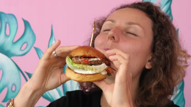 A very hungry young woman is eating her vegan burger with great pleasure and joy. She holds it with both hands, throwing her head back with her mouth wide open, she bites off a piece.