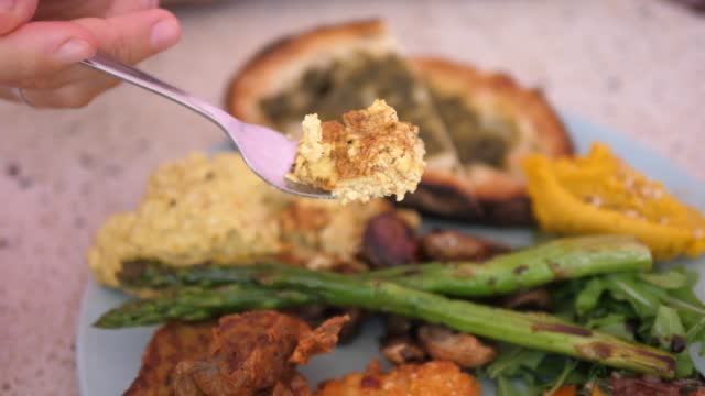 Close-up of a vegan omelet dinner with tofu, asparagus, hummus, toast and grilled vegetables. The hand breaks the omelet and the piece is brought closer to the camera.