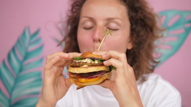 Close-up of a hungry caucasian woman. She holds a two-tiered vegan burger in both hands, then she takes a bite of it while her eyes close in pleasure.