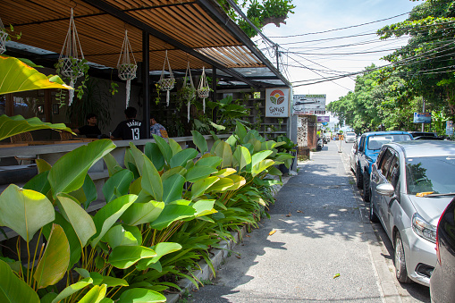 Sanur, Bali, Indonesia - March 14, 2022.
View of Jl. Hang Tuah in north Sanur, Bali, Indonesia with Canvas Cafe on the left side and lots of tropical plants in front and parked cars on the right side. Taken during the coronavirus lockdowns and with no tourists in sight.