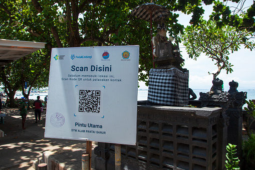 Sanur, Bali, Indonesia - March 14, 2022.\nA large sign at north Sanur Beach near Sanur Port, Bali, Indonesia, asking people to scan their QR codes on their PeduliLindungi App in order to enter this part of Sanur. Translated the sign says 'Scan Here' and 'Main Entrance'. A Hindu statue can be seen behind the sign and the sea just beyond.