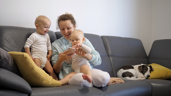 Happy family concept. A young mother plays with her curious twin girls by showing them something on her smartphone. A cute dog sleeps on the couch next to them.