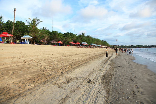 kuta beach à bali, en indonésie, avec du sable blanc, des parasols à la limite des arbres et des gens marchant le long de la plage avec un fond de ciel bleu. - parasol umbrella asian ethnicity asian culture photos et images de collection