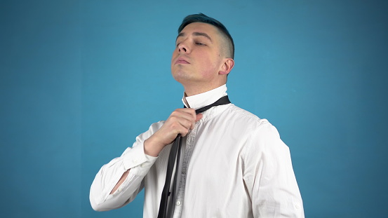 A young man in glasses with blue hair is dressing up a suit. An alternative person fastens buttons and puts a tie on himself on a blue background. 4k