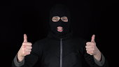 A man in a balaclava mask shows his approval mark with his hands. The thug raised his hands and showed his thumbs up against a black background.
