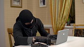 A robber is trying to hack into a laptop. A masked thug is sitting in a house and trying to break into a laptop. Theft of data from a computer.