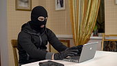 A robber is trying to hack into a laptop. A masked thug is sitting in a house and cannot hack into a laptop. Theft of data from a computer.