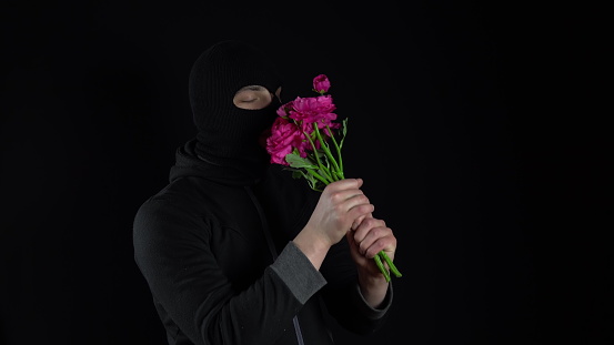 A man in a balaclava mask is standing with flowers. Bandit sniffs a bouquet of pink flowers on a black background. 4k