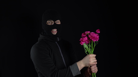 A man in a balaclava mask is standing with flowers. Bandit sniffs a bouquet of pink flowers on a black background. 4k