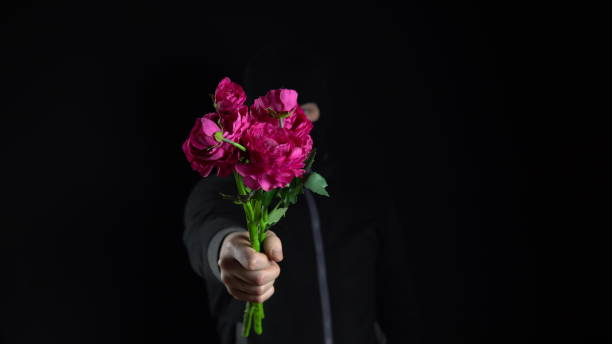 a man in a balaclava mask is standing with flowers. the bandit holds out a bouquet of pink flowers to the camera. on a black background. - convict lake imagens e fotografias de stock