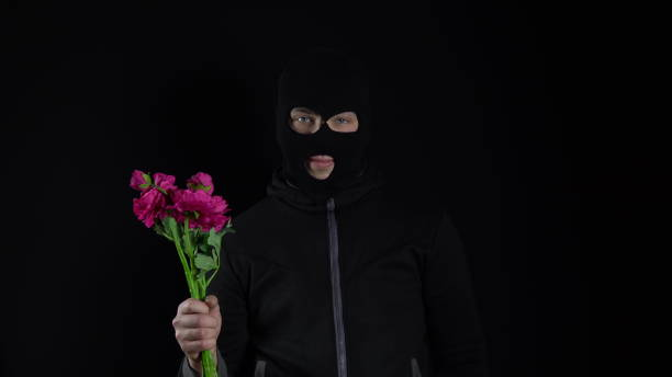 a man in a balaclava mask is standing with flowers. the bandit holds in his hand a bouquet of pink flowers and smiles on a black background. - convict lake imagens e fotografias de stock
