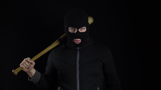 A man in a balaclava mask is standing with a baseball bat. A bandit stands on a black background with a baseball bat over his shoulder. 4k