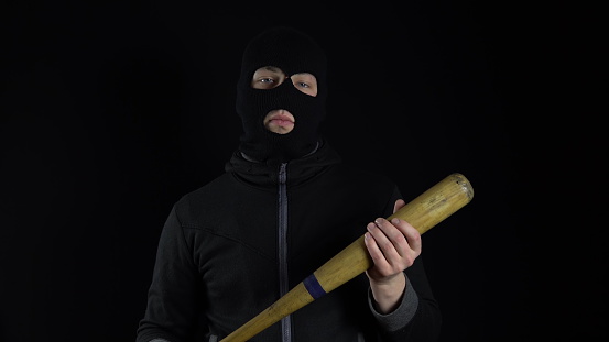 A man in a balaclava mask is standing with a baseball bat. A gangster is holding a baseball bat on a black background. 4k
