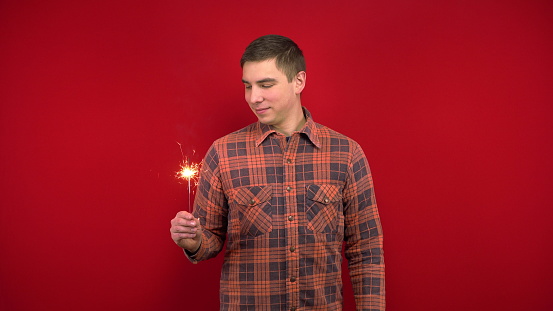 A young man in a red shirt holds a sparkler in his hand. Shooting in the studio on a red background. 4k