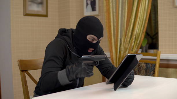 istock A robber is trying to hack into a tablet. A masked thug is sitting in a house trying to break into a tablet and is threatening with a gun. Theft of data from a tablet. 1444599293